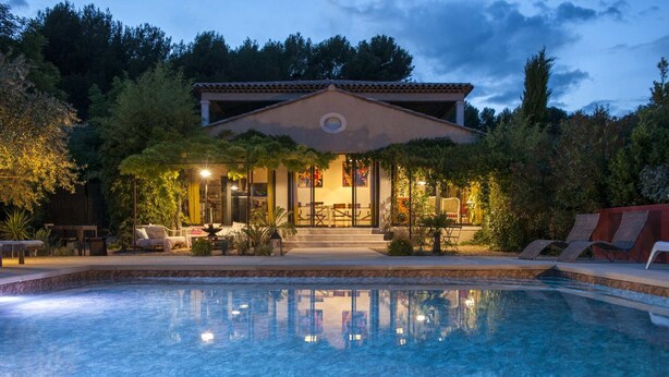 Un espace aménagé autour d’une piscine, avec une maison en arrière-plan, le tout entouré d’arbres et sous un ciel dégagé.