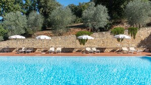Une piscine entourée de murs de pierre et d’arbres, avec des chaises longues et des parasols.