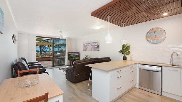 A modern kitchen with a built-in dishwasher, a dining area with a wooden table, and a living room with a sofa and TV.