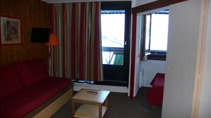 A cozy room with a red sofa, a small wooden table, and a window with a view of a deck and mountains. - Reberty les Menuires very beautiful 2 rooms facing south (saint martin de belleville)