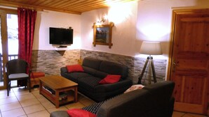 Una sala de estar con un sofá, una butaca, una mesita, un televisor montado en la pared y una lámpara.
