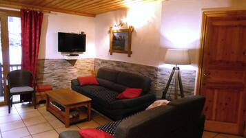 Un salon comprenant un canapé, un fauteuil, une petite table, un téléviseur fixé au mur et une lampe.