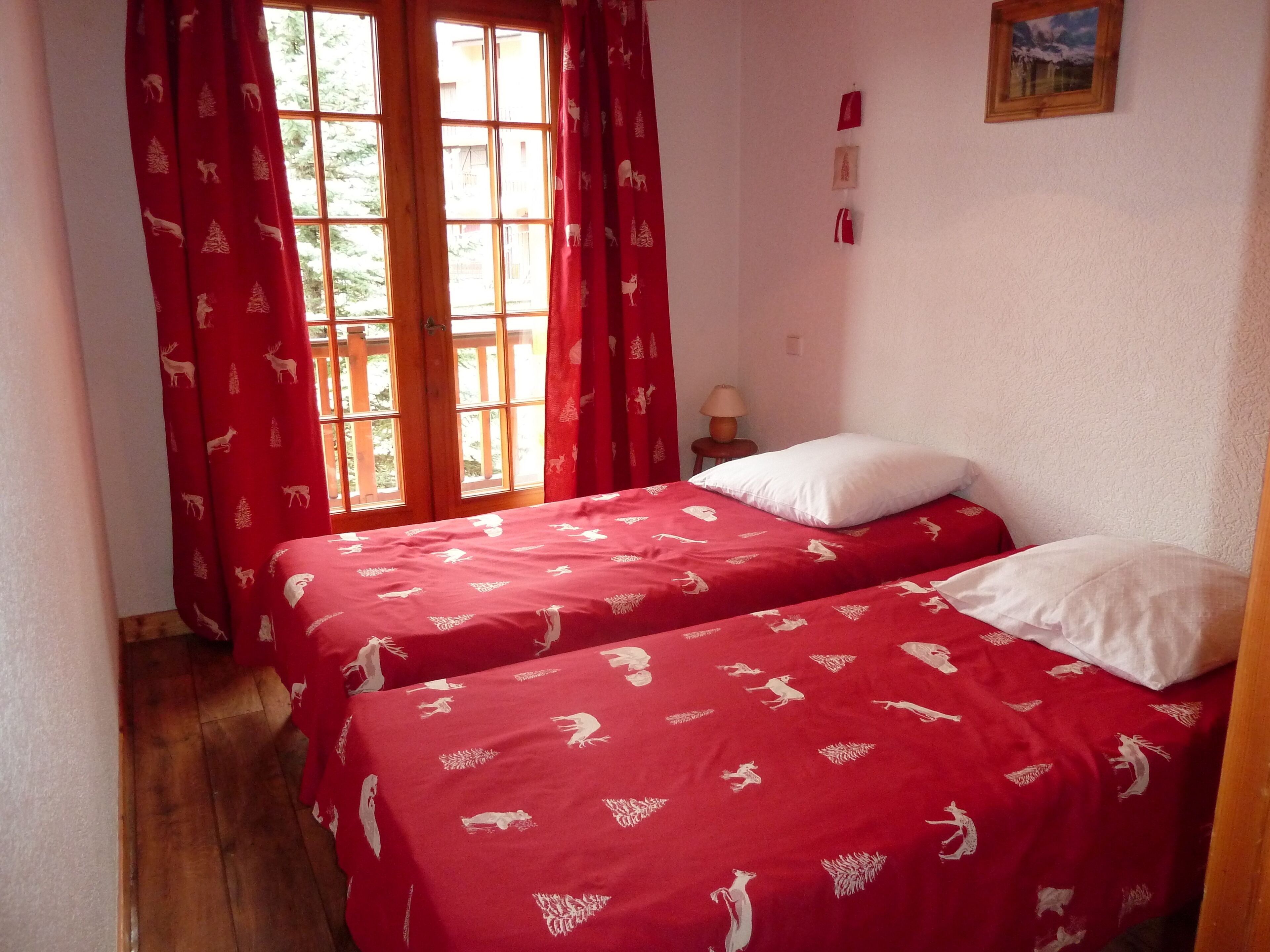 Une chambre avec deux lits, des rideaux rouges à motifs animaliers, un plancher en bois, une petite table avec une lampe et un tableau encadré au mur.