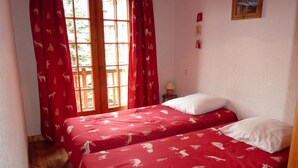 Una habitación con dos camas, cortinas rojas con estampados de animales, piso de madera, una mesita con una lámpara y un cuadro en la pared.