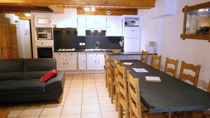 Una sala de estar acogedora con un sofá oscuro y una mesa de comedor con sillas de madera. Se ve una cocina con armarios blancos y baldosas negras al fondo.