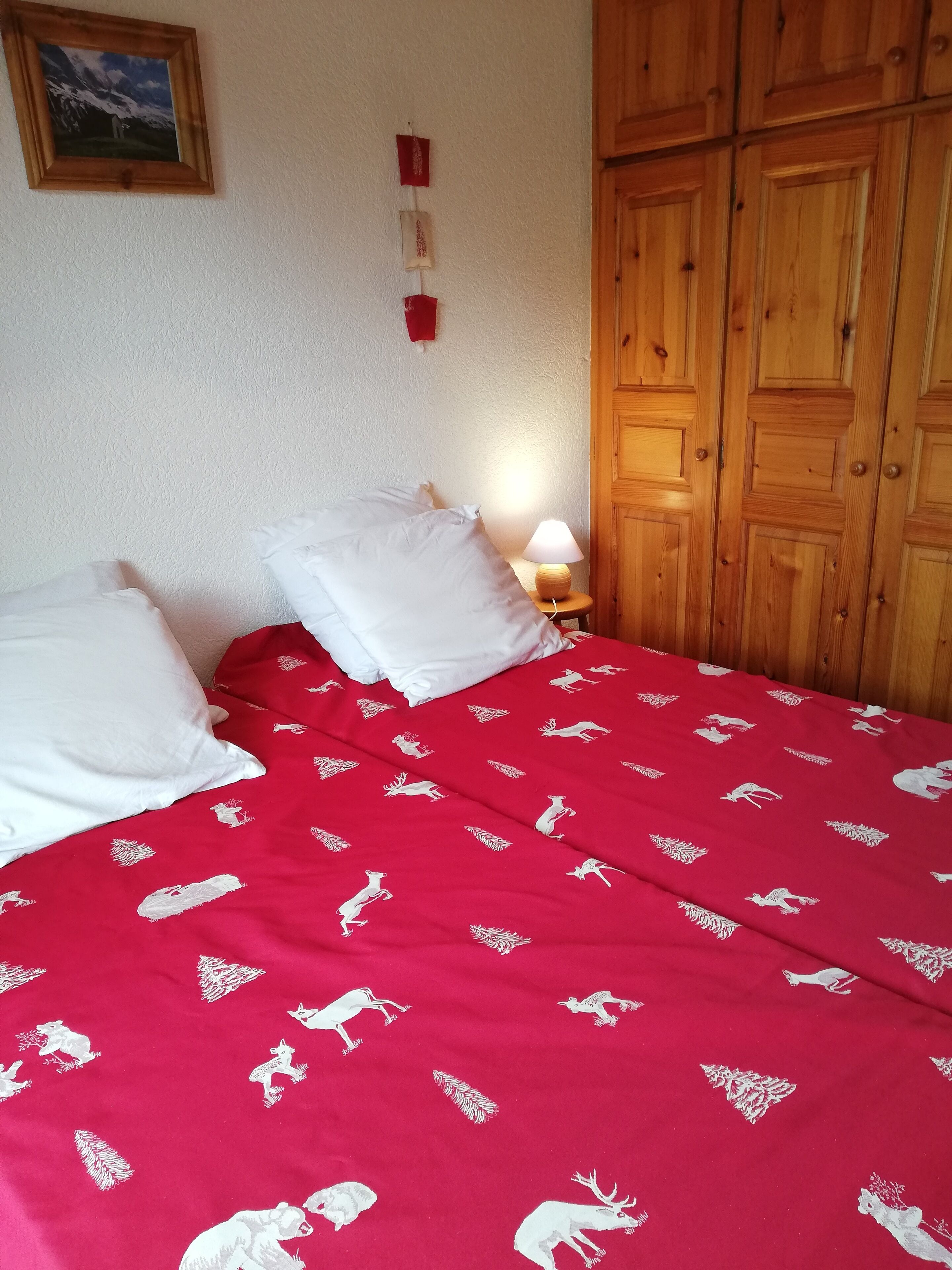 A bedroom with a red bedspread featuring animal prints, a wooden wardrobe, and a small bedside table with a lamp.