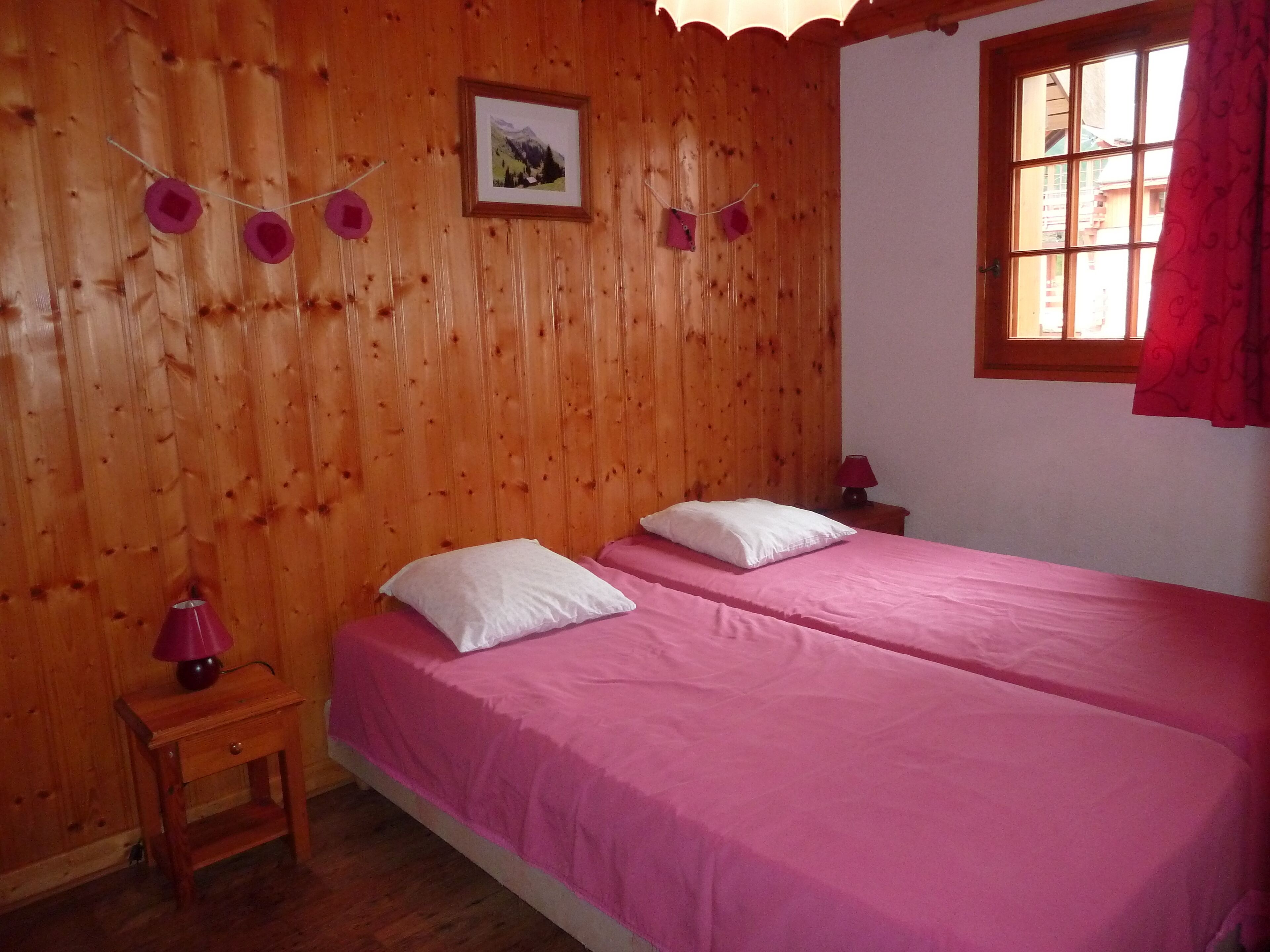 Una habitación con paredes de madera, dos camas con ropa de cama rosa, una mesita de noche de madera con una lámpara y una ventana con cortina.