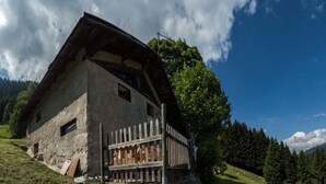 Ein rustikales Steinhaus mit geneigtem Dach, Holzzaun und Blick auf ein bewaldetes Gebirge.