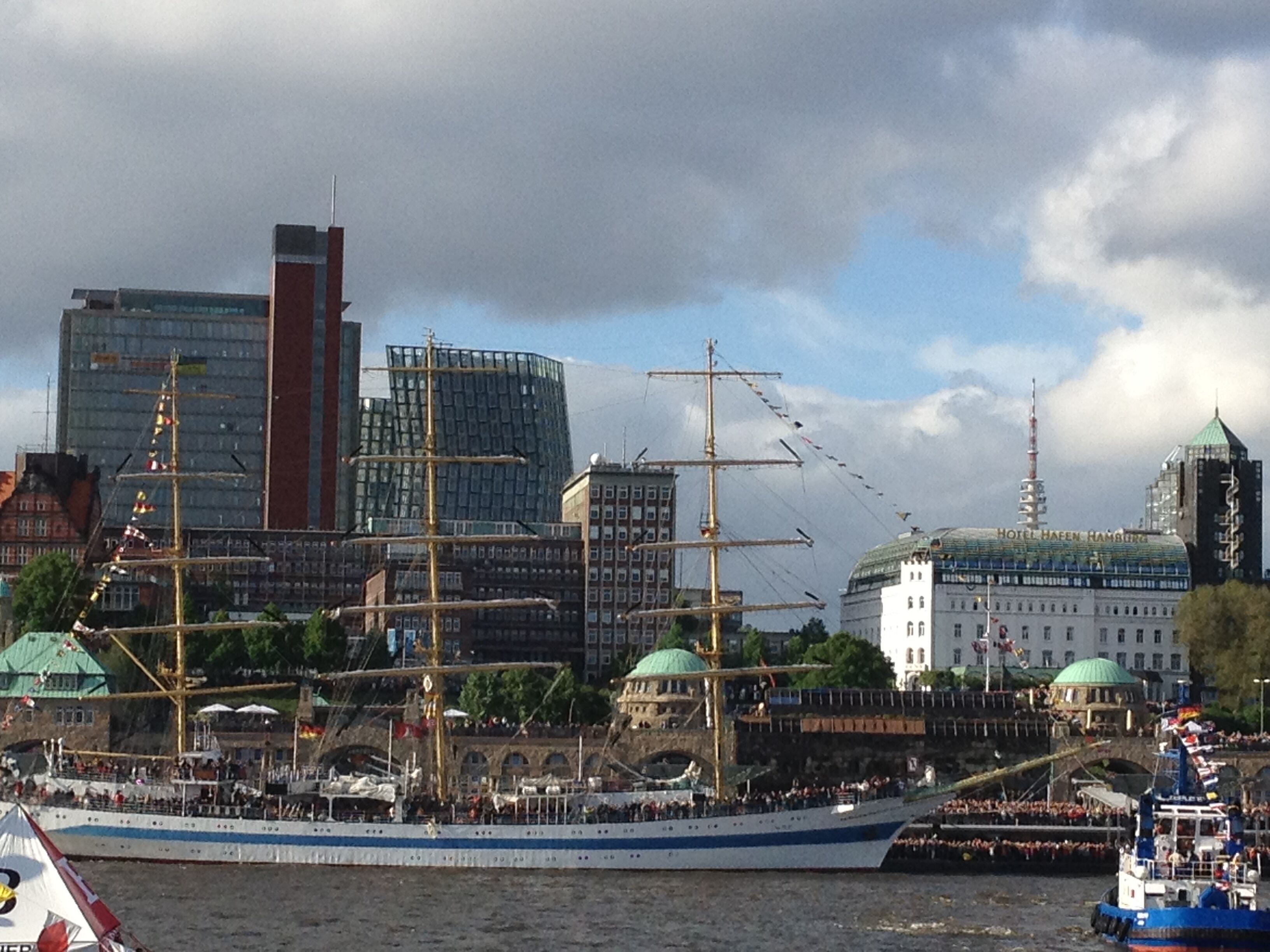 A large sailing ship docked at a harbor with a city skyline in the background.