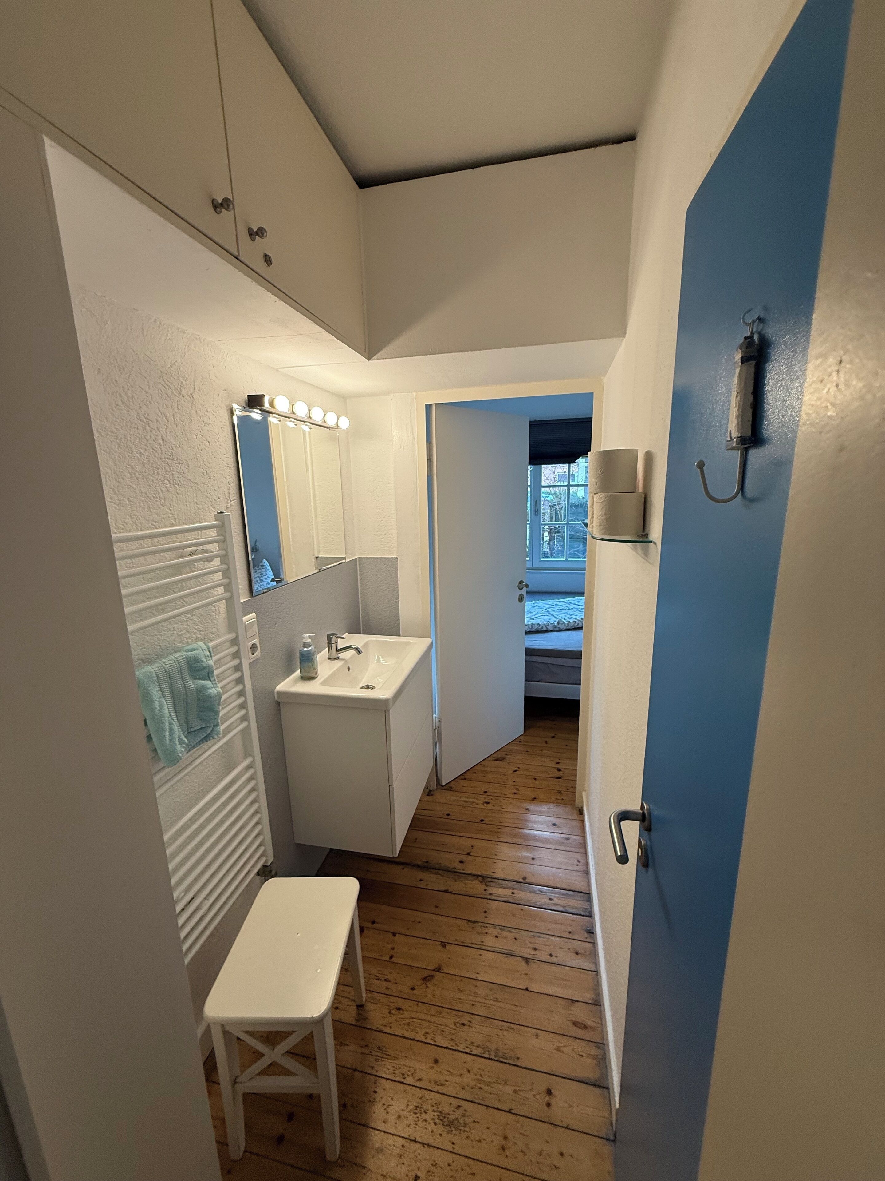 Un couloir étroit avec un lavabo blanc, un miroir avec des lumières, une porte bleue et un plancher en bois.
