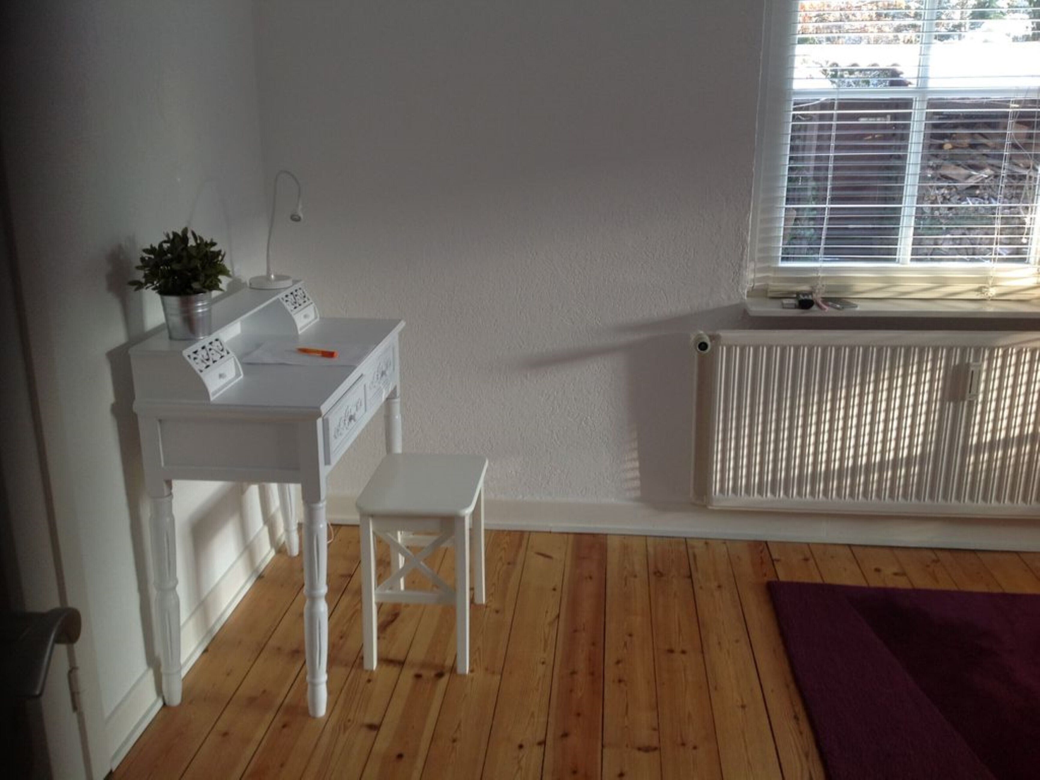 A small, tidy room with a white desk, a chair, a radiator, and a window with blinds.