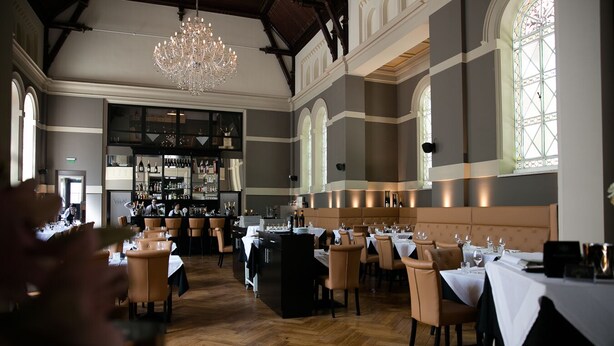 A restaurant with high ceilings, chandeliers, and a bar area. Tables are set with white tablecloths and wine glasses.