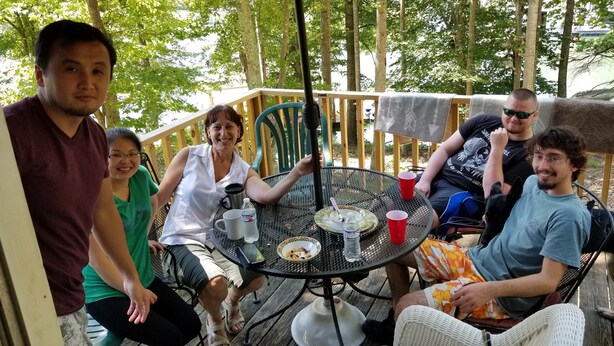Un groupe de personnes est assis autour d’une table ronde en métal, avec des chaises, une terrasse en bois et une rambarde en arrière-plan.