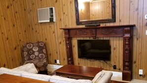Un salon avec des murs lambrissés, un grand téléviseur à écran plat fixé au-dessus d’une cheminée en bois, un fauteuil à motifs et un ensemble de canapés.