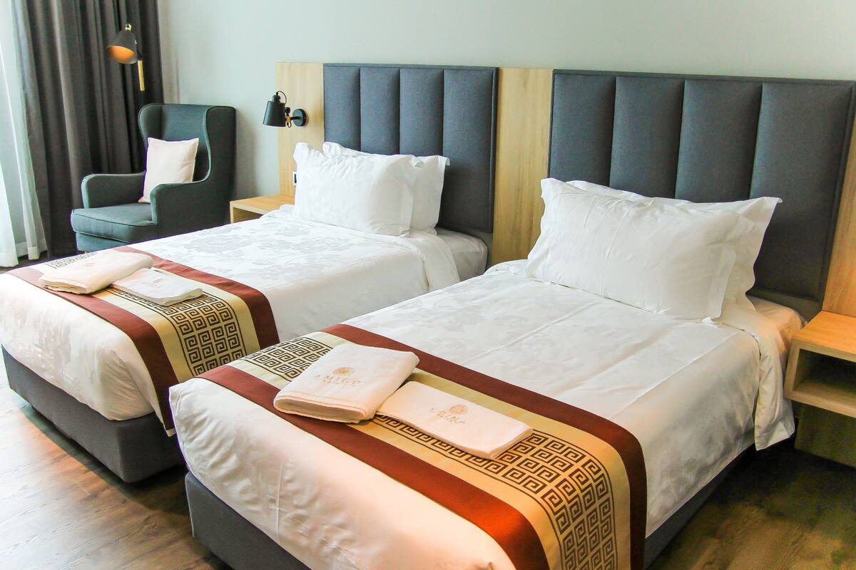 A hotel room with two beds, each with white linens and decorative pillows. There is a chair and a nightstand with a lamp.