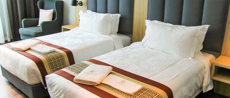 A hotel room with two beds, each with white linens and decorative pillows. There is a chair and a nightstand with a lamp.