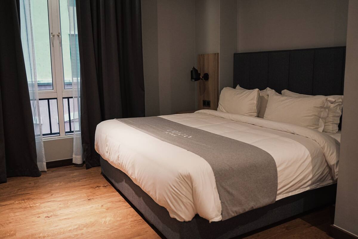A neatly made bed with a grey and white duvet, a wooden headboard, and a nightstand with a lamp.