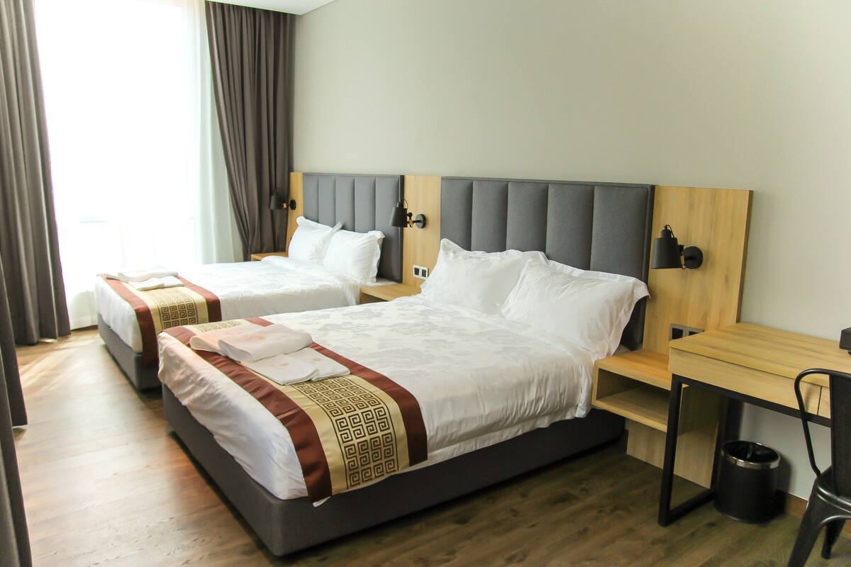 A hotel room with two beds, a wooden desk, and a trash can.