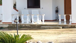 A white building with a porch, two white chairs, a potted plant, and a wooden door.