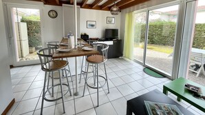 A dining area with a wooden table and bar stools, a kitchen area with a TV and counter, and large sliding glass doors to a garden. - Semi-detached bungalow near Lac Nord (067) (Biscarrosse)
