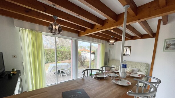A modern kitchen with wooden beams, a dining area with a table set for four, and a sliding glass door leading to a garden. - Semi-detached bungalow near Lac Nord (067) (Biscarrosse)