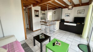A living room with a sofa, a coffee table, and a TV. There is a kitchen area with a dining table and chairs. The room has wooden beams on the ceiling. - Semi-detached bungalow near Lac Nord (067) (Biscarrosse)