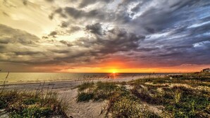 Een strand met een zonsondergang, zandduinen en een bewolkte lucht.