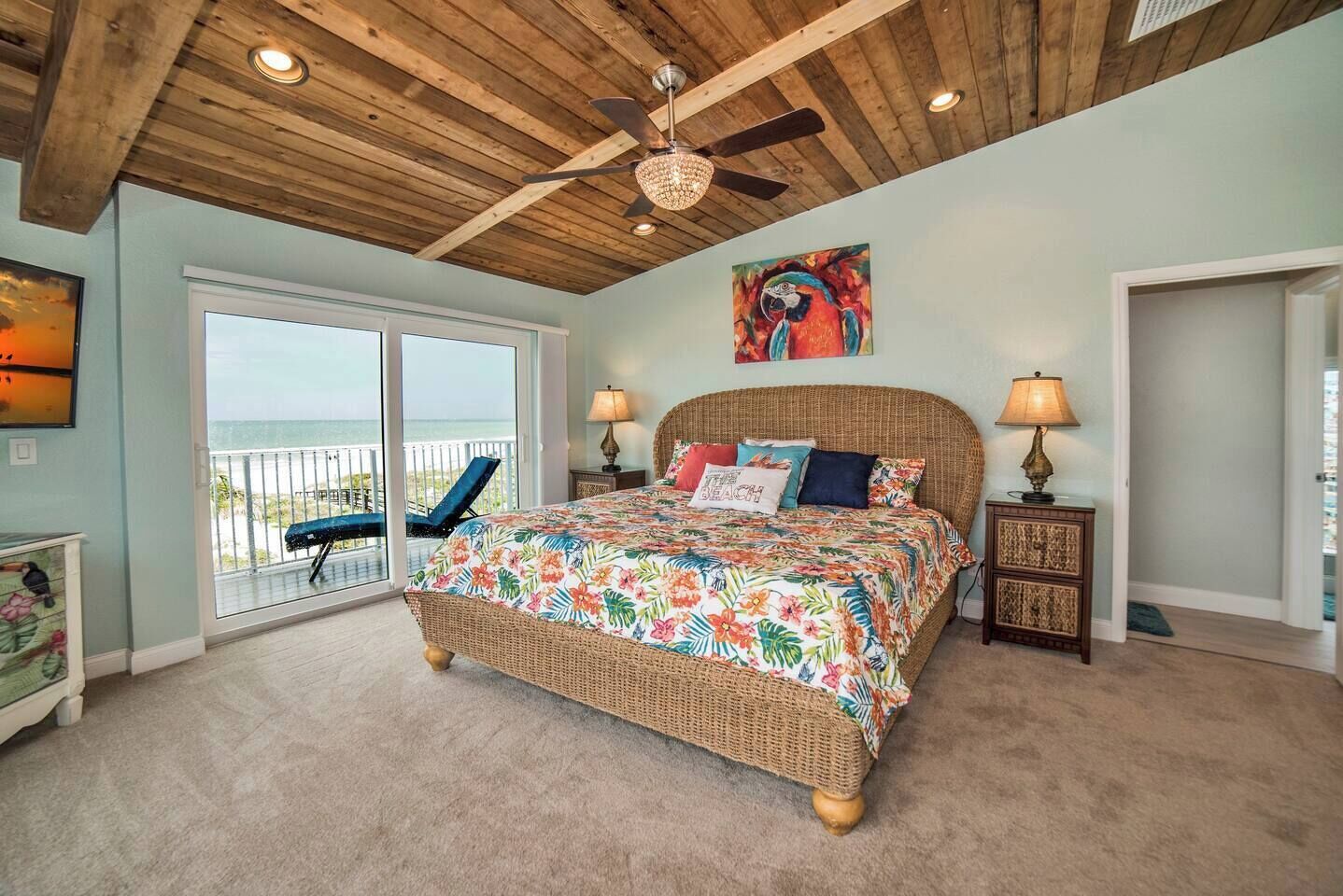 A bedroom with a large bed, a ceiling fan, a painting, and a view of the ocean through a sliding door.