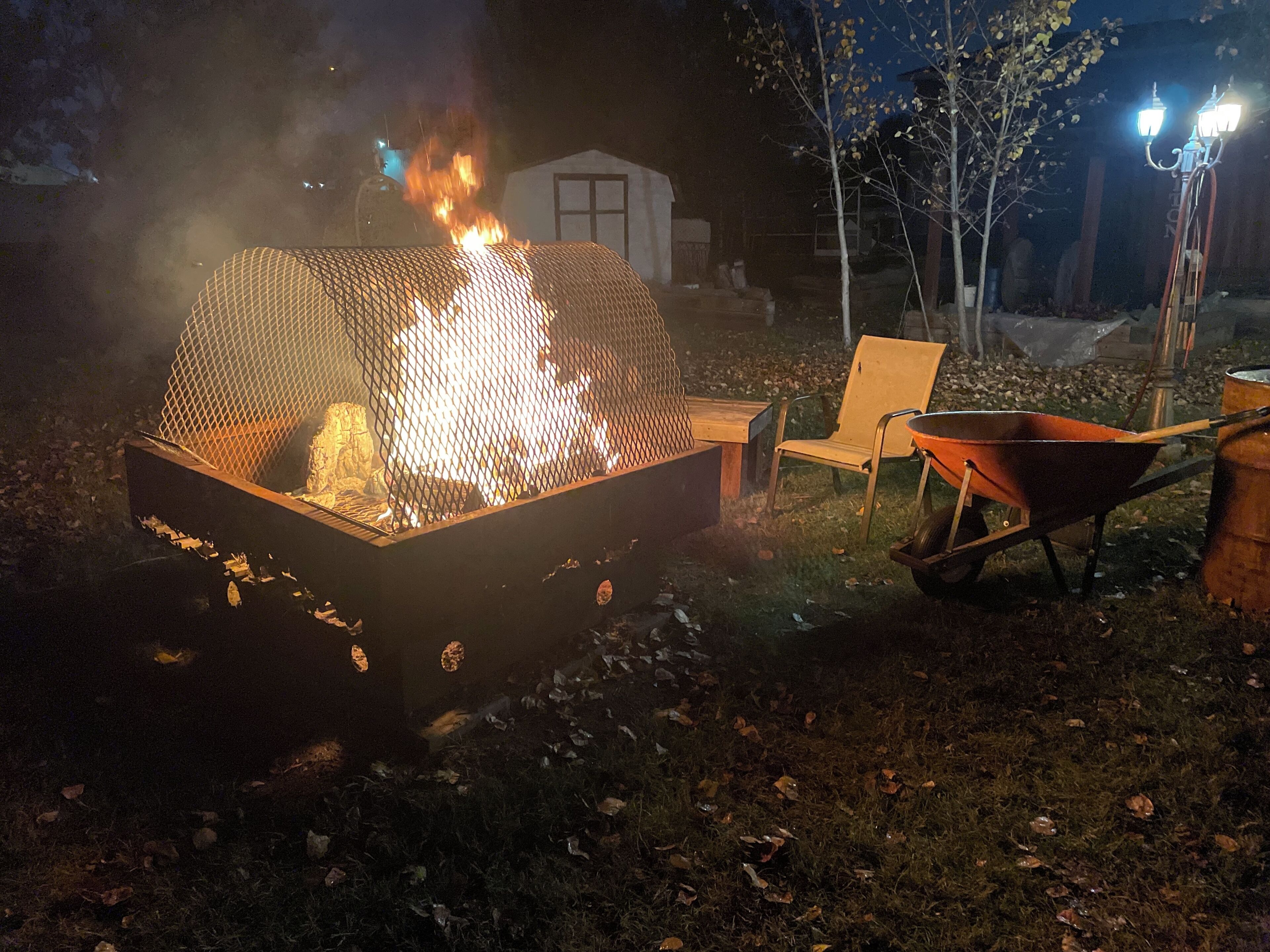 A fire pit with a mesh cover, a wheelbarrow, and a chair in a backyard at night.