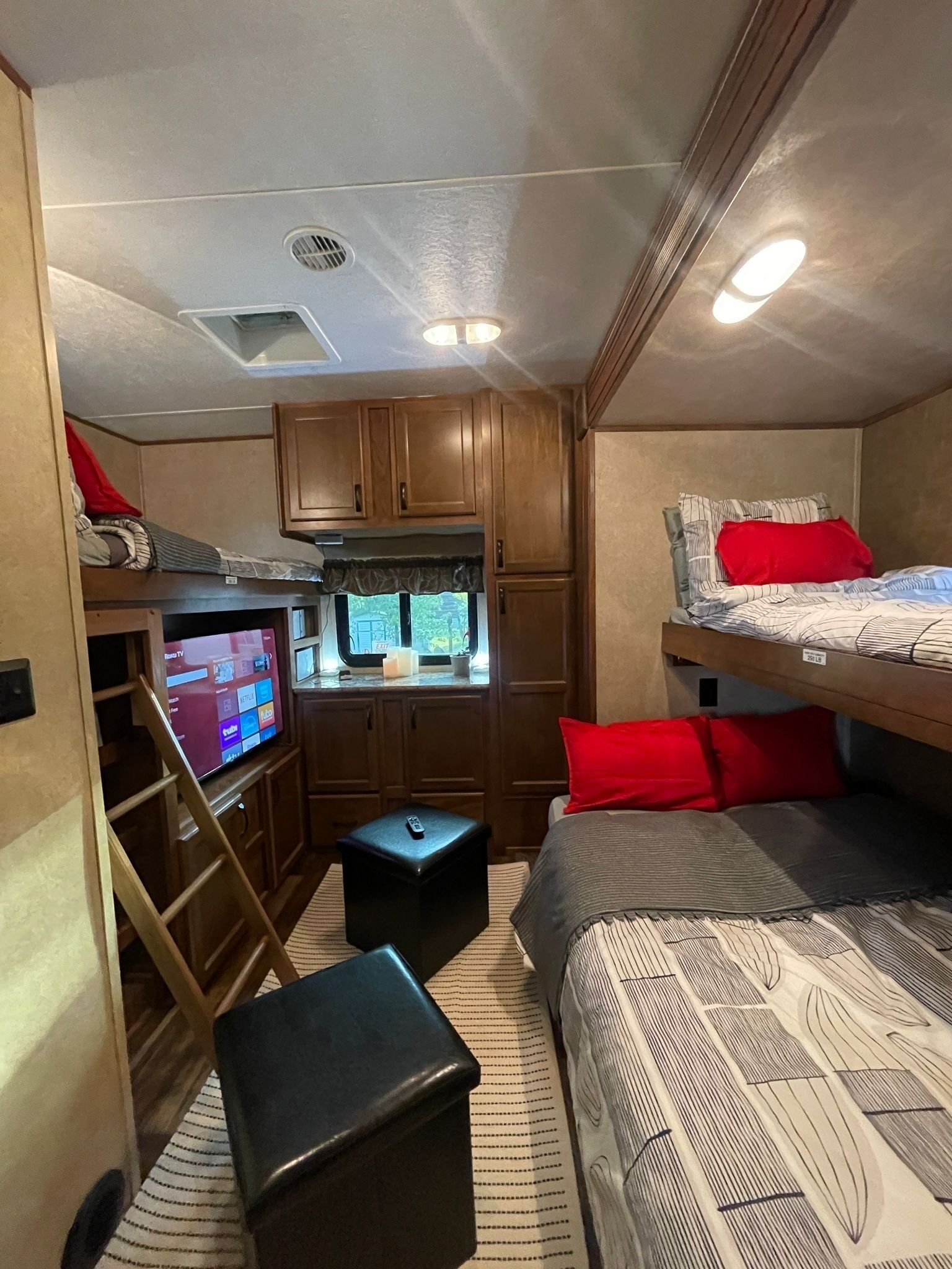 Interior compacto de una autocaravana con kitchenette, televisor, literas y un pequeño reposapies con almacenamiento.
