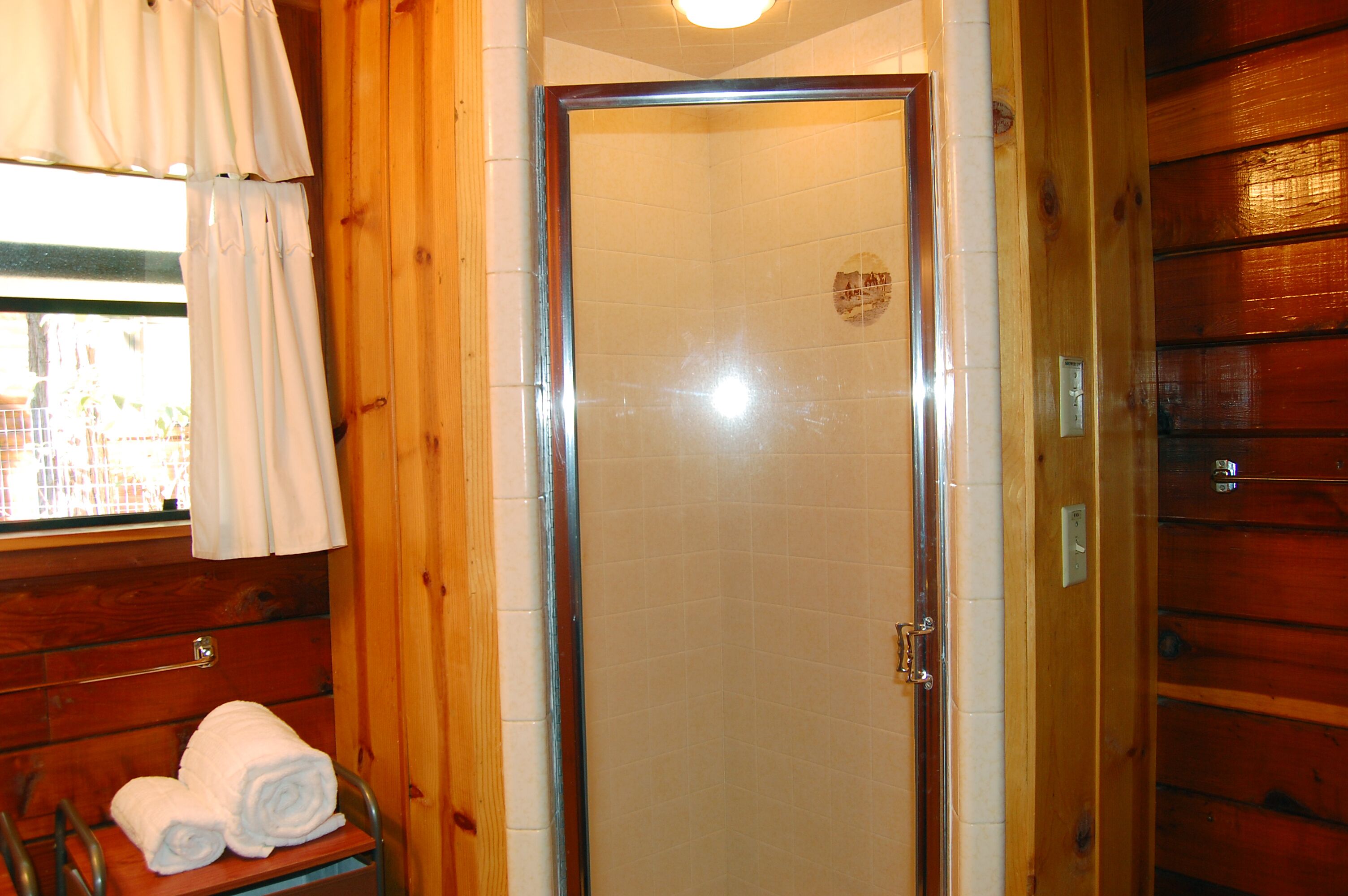 Baño de cabaña de madera con mampara de ducha, toallero con toallas y ventana con cortinas.