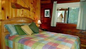 A wooden cabin bedroom with a bed, colorful quilt, wooden headboard, and a window with curtains.