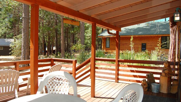 A wooden deck with white chairs, a covered area with a wooden railing, and a cabin in the background.