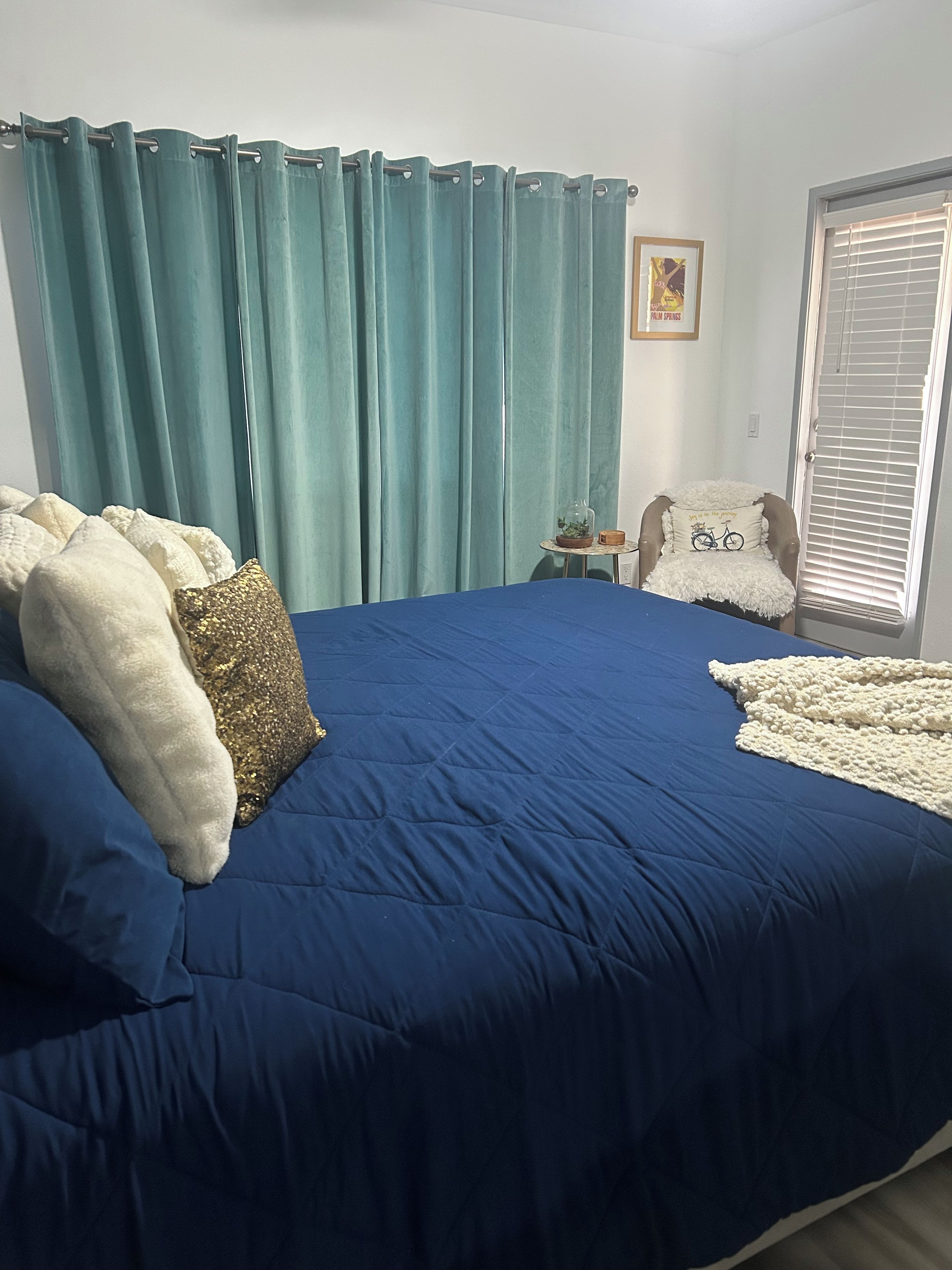 Quarto com uma cama grande, cortinas verde-água, uma mesinha de cabeceira com abajur e uma cadeira com almofada.