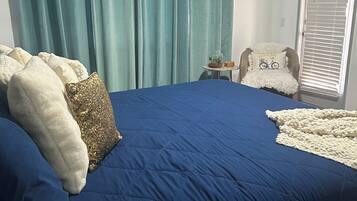 Quarto com uma cama grande, cortinas verde-água, uma mesinha de cabeceira com abajur e uma cadeira com almofada.