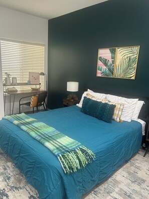 Een slaapkamer met een groot bed, een bureau met stoel, een lamp en een lijst met afbeelding aan de muur.
