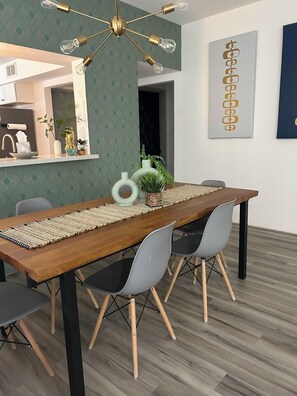 A dining area with a wooden table, grey chairs, a woven placemat, and a modern chandelier. - Palm Desert Sunshine Escape - Heated Pool & Fall Getaway (Palm Desert)