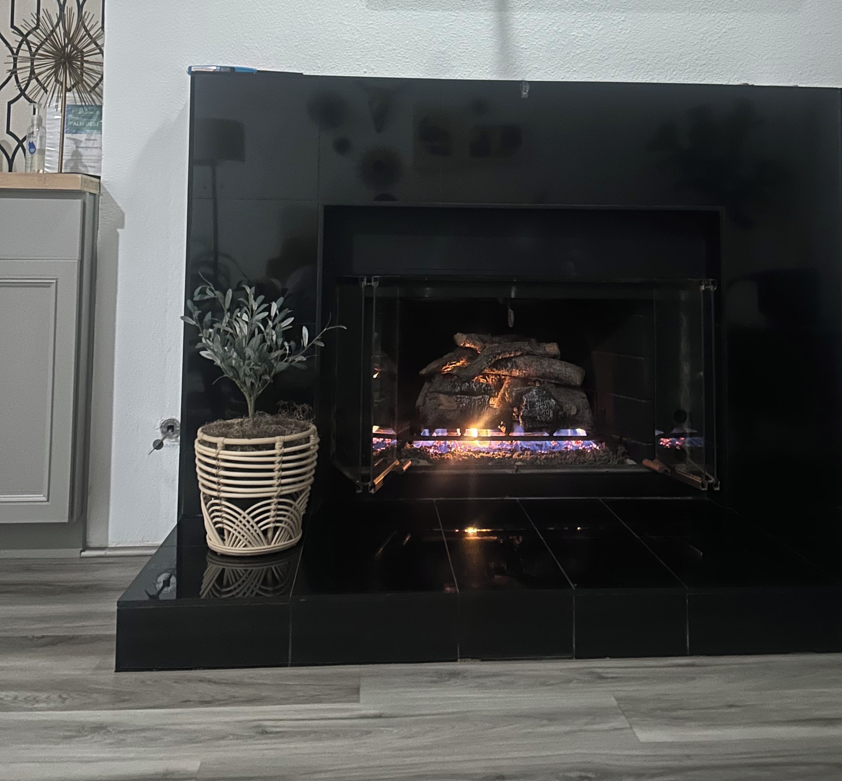 Una chimenea con fuego de leña, una cesta de mimbre con una planta y una puerta blanca al fondo.