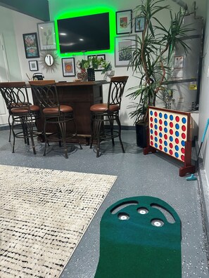 Un comedor con barra, sillas, alfombra verde y máquina de pinball.