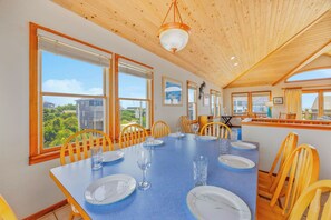 Comedor con una mesa larga dispuesta para seis personas, sillas de madera, ventanas amplias con vistas a zonas verdes y edificios, y techo de madera.