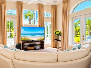 A living room with a large flat-screen TV, beige curtains, and a view of a tropical landscape.