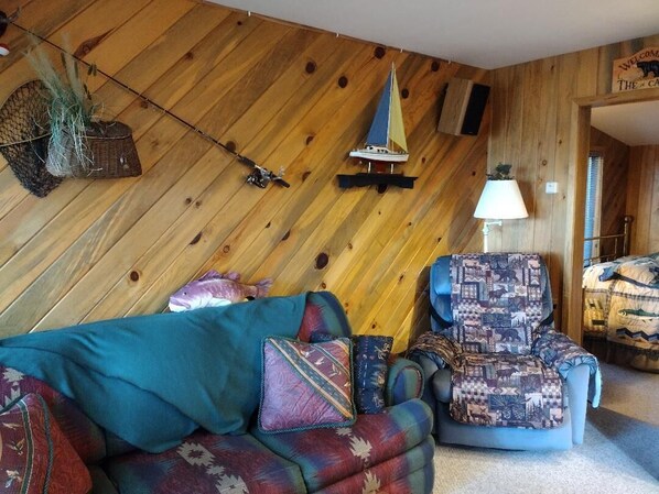 Una habitación acogedora con pared de madera, un sofá, un sillón estampado, un pequeño modelo de velero y un cartel de bienvenida.