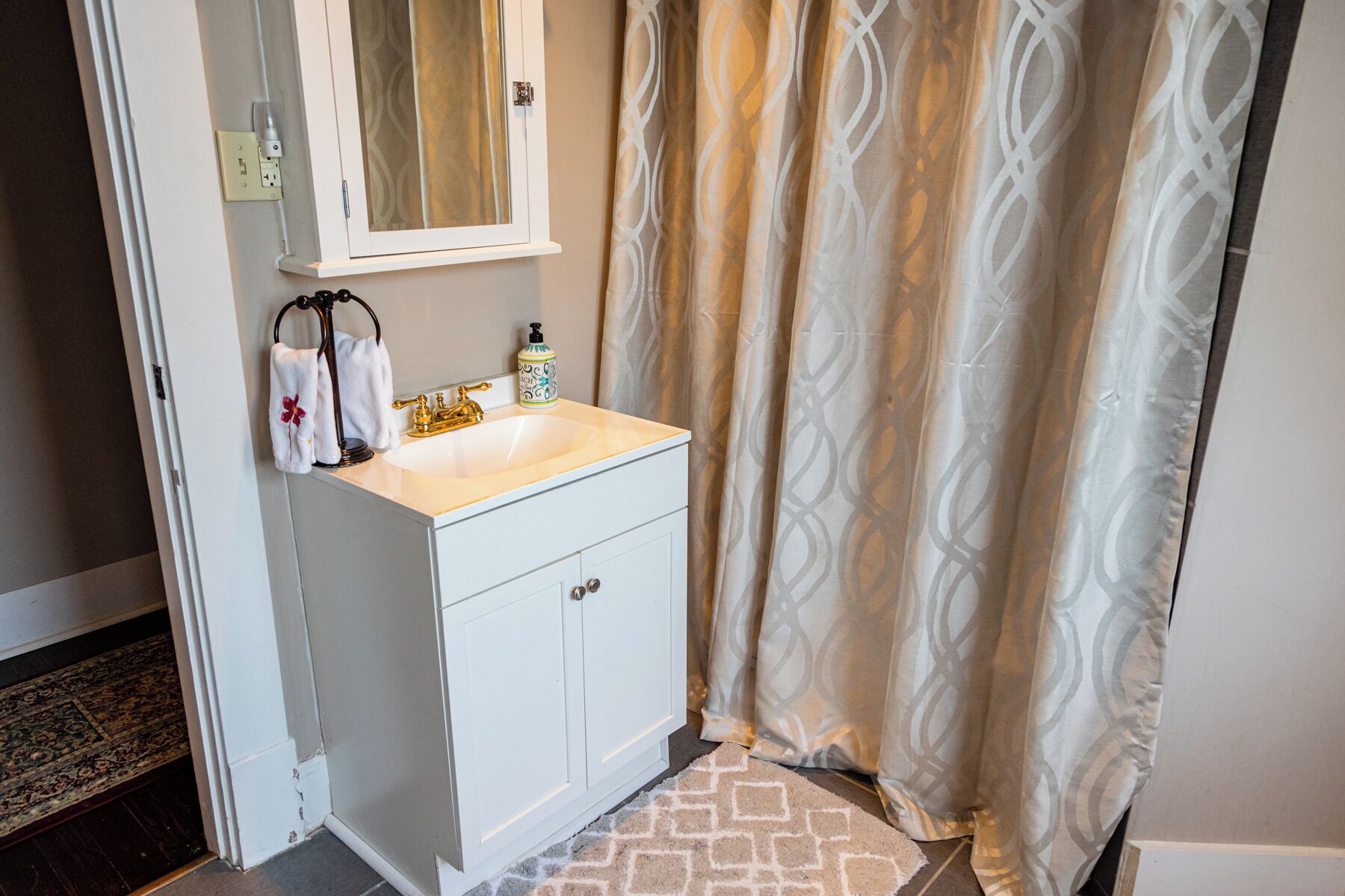 A bathroom with a white vanity, a gold faucet, a bottle of liquid soap, and a patterned shower curtain.