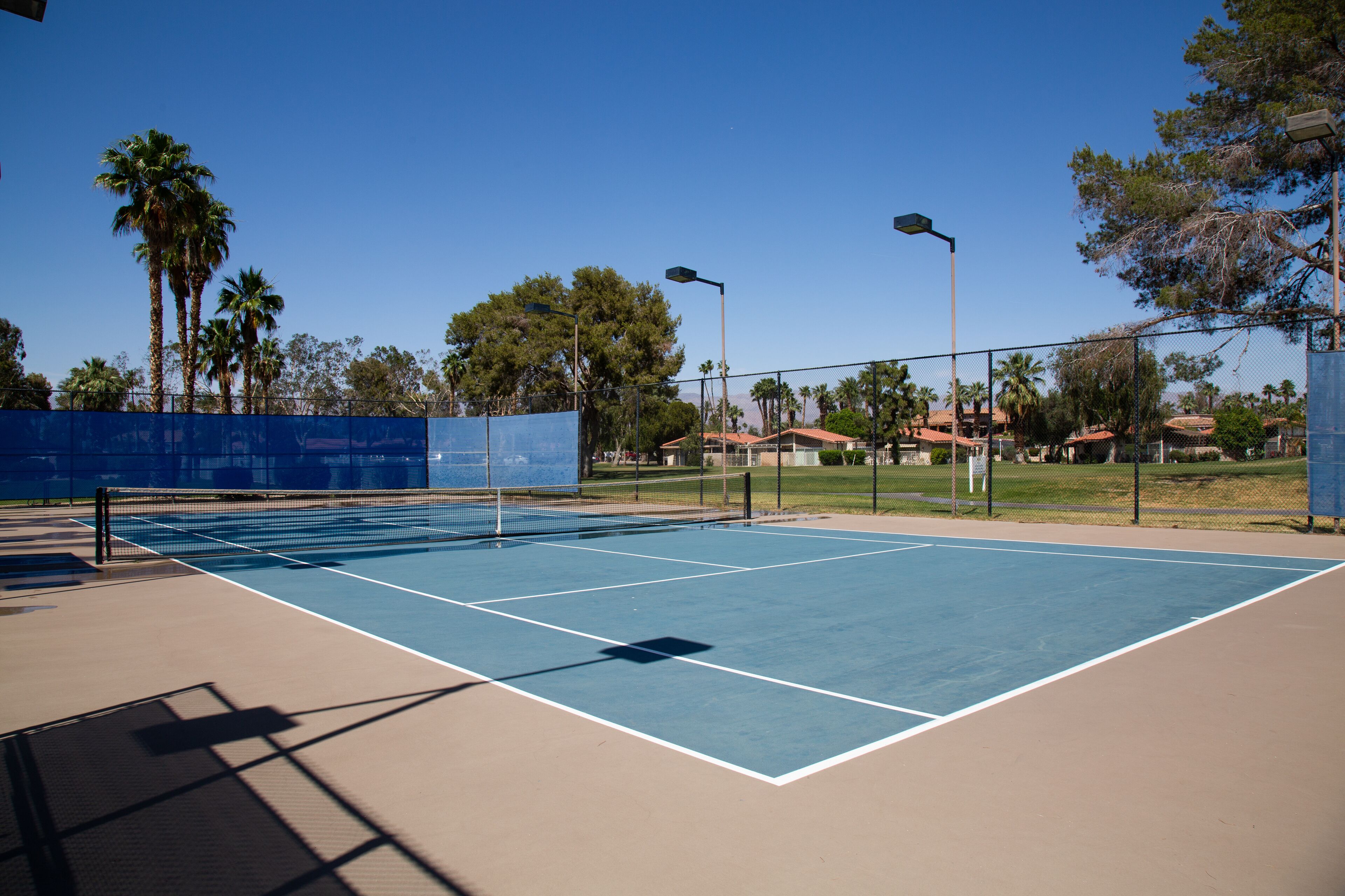 Un court de tennis en plein air, doté d’une surface bleue, entouré d’une clôture et de palmiers.