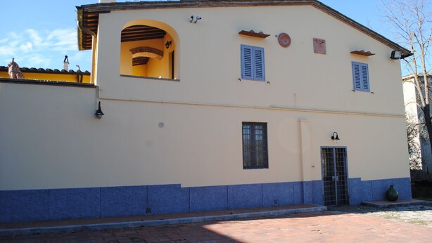 A two-story building with a yellow facade and blue trim, featuring a central arched doorway, a small window with shutters, and a door with a metal gate.