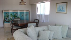 Un salon comprenant un canapé, une table à manger avec des chaises, un paravent décoratif orné de motifs d’oiseaux et une fenêtre avec des rideaux.