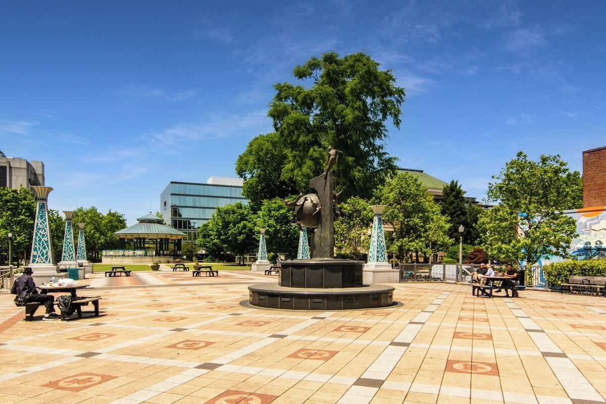 Une place publique agrémentée d’une sculpture centrale, de bancs et d’arbres.