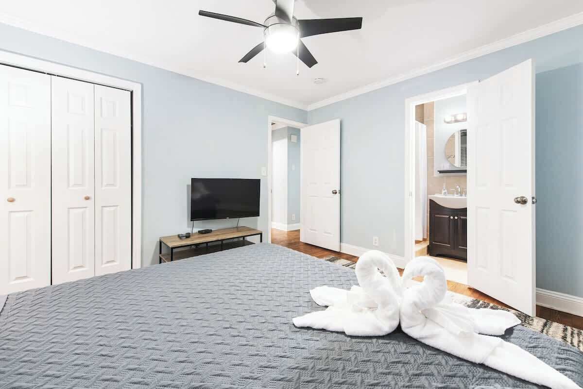 Une chambre à coucher avec un lit, un ventilateur de plafond, un téléviseur et une salle de bain visible à travers une porte ouverte.