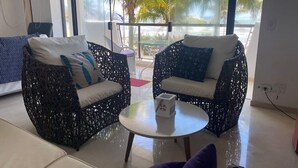 Quarto com duas cadeiras de vime, uma mesa redonda e vista para palmeiras através de amplas janelas.
