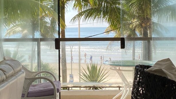 Quarto com vista para a praia, um sofá de vime, uma mesa de vidro e uma cadeira de fibra preta.